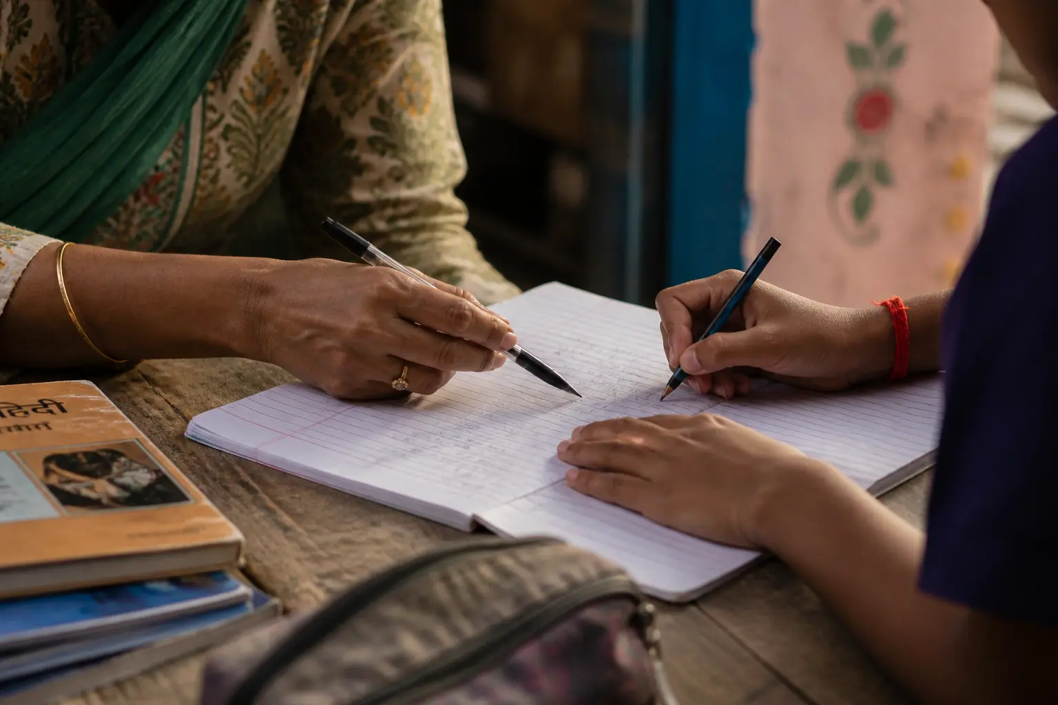 Mentor guiding a student while writing in a notebook, close-up of hands, real Indian learning environment